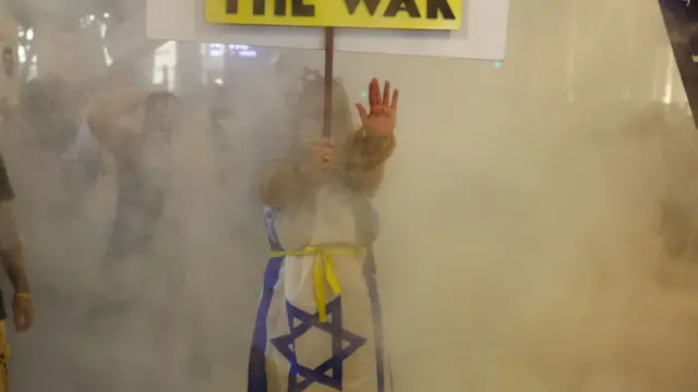 A woman holds a protest sign and her hand up, palm facing outwards, with ropes around her wrists. She wears a dress with the Israeli flag on it and a yellow ribbon symbolising the hostages around her waist.
