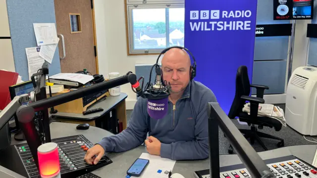 Ben sits in a radio studio, in front of a microphone and controls. He wears a blue fleece and headphones, and has a serious expression.
