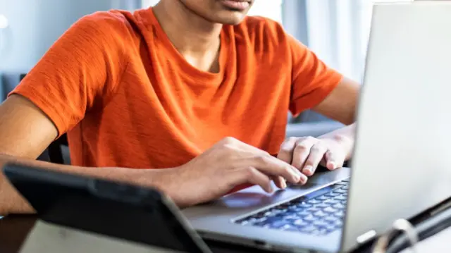 A cropped image of a boy in an orange t-shirt, using a laptop.