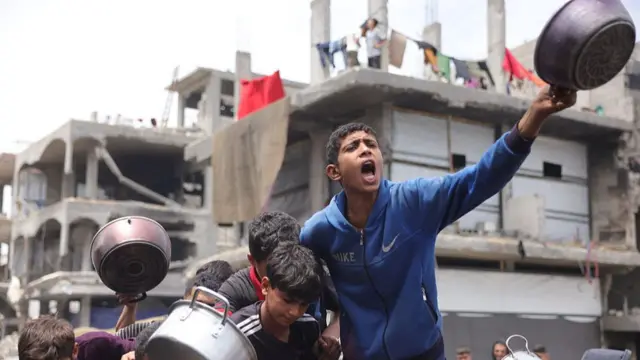 A young boy in a blue jacket holds up a silver pan in front of destroyed buildings in Gaza