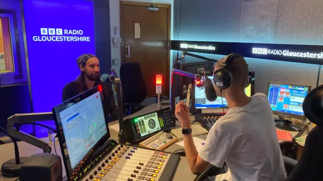 BBC Radio Gloucestershire presenter Jon Smith sits in the studio with John De Gruyther. They are facing each other, and there are lots of screens and boards with controls on them.