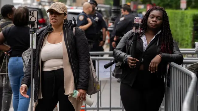 Streamers produce content outside the federal court. One woman is seen speaking into the camera of her phone that is mounted on a tripod. Another woman is seen holding her small black tripod on which a phone is mounted