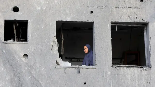A Palestinian in a hijab looks out the window with no glass of a partially destroyed building