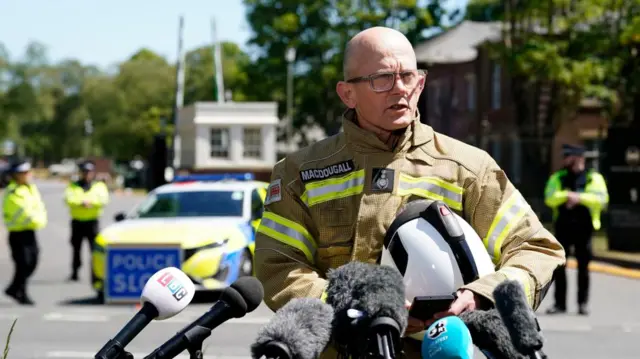 Oxfordshire Fire and Rescue Service Chief Fire Officer, Rob MacDougall, speaks to the media. He is wearing a beige and fluorescent yellow firemans uniform and carrying a helmet.