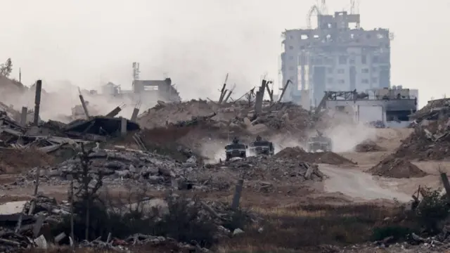 Three Israeli military Humvee trucks driving near the Gaza border, there is much rubble from destroyed builings around them and a heavily damaged high rise building in the backdrop