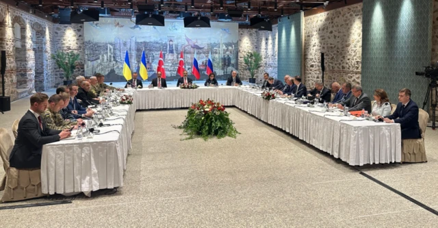 A large table with people sat on chairs with microphones, Ukraine, Turkey and Russia flags behind them.