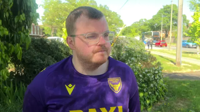 A man with short blonde hair, glasses and wearing a purple and yellow Oxford United t-shirt looking at something off-camera. There's a street and shrubbery behind him and the sun is out.