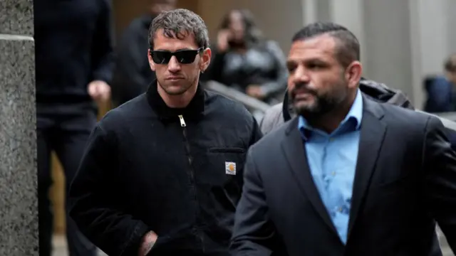 A man in dark clothing and sunglasses walks behind a man in a suit.
