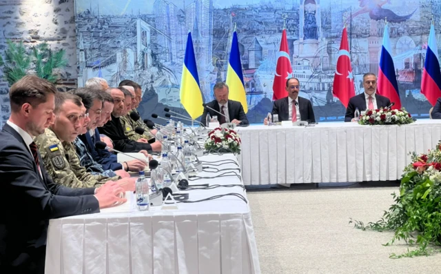 Row of men sat at a table with Ukraine's flag behind them.