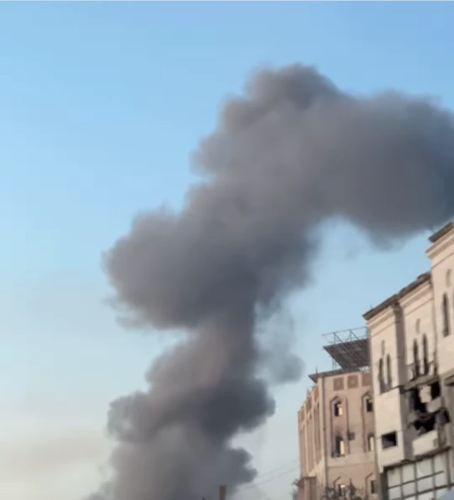A screengrab from a video showing a thick plume of black smoke rising into the air. A heavily damaged building is visible to its right.