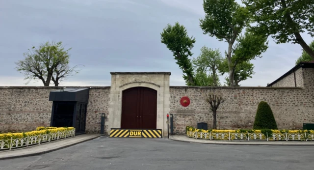 A wall with a large door in the middle, which is closed, trees and shrubs nearby and a large black and yellow barrier saying DUR