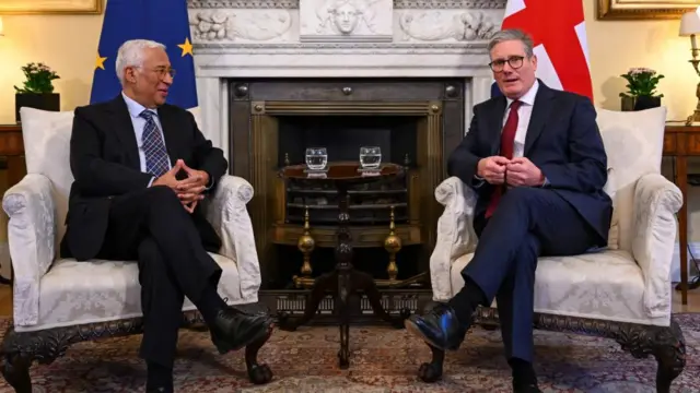 Antonio Costa and Kei Starmer sat on chairs with EU and UK flags behind them. A table stands in between the chairs with two glasses of water on it.