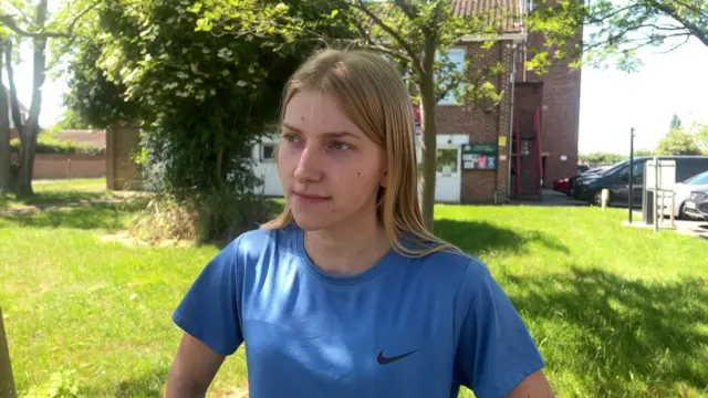 A young woman with long blonde hair and wearing a blue T-shirt looking at something off-camera. There's a small stretch of grass, some trees and a brick building behind her.