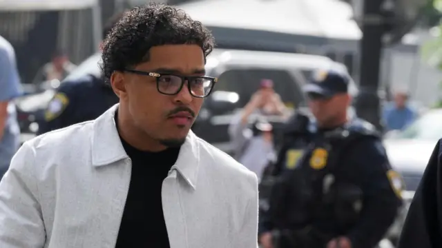 A man wearing glasses and white jacket walks past a police officer.