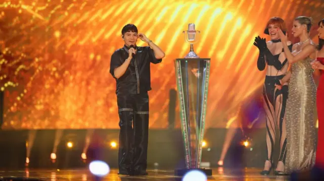 JJ on stage next to a podium holding up the crystal mic award as Nemo, Michelle Hunziker and Sandra Suder applaud him from the sidelines