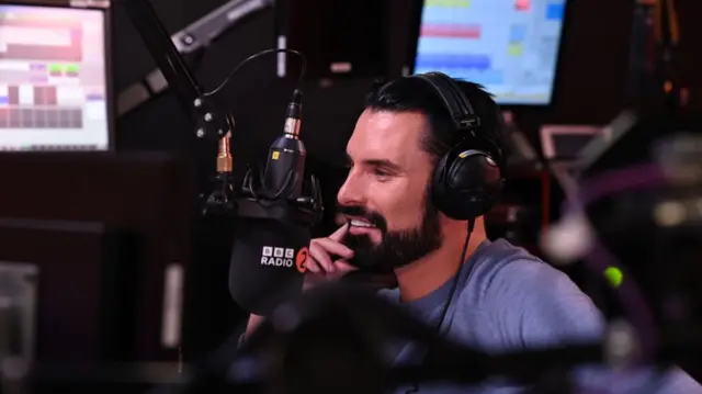 Rylan Clark in a blue long-sleeved shirt sits in a radio studio surrounded by broadcasting equipment, including a black mic with the BBC logo and two monitors