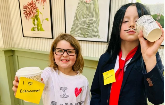 Finn (L: girl with light brown hair, black glasses holding a take away coffee cup with a yellow post-it reading I love Eurovision) and Charlie (R, wearing a straight black hair wig, black blazer and red tie as he pretends to drink from a paper cup) in Eurovision costumes