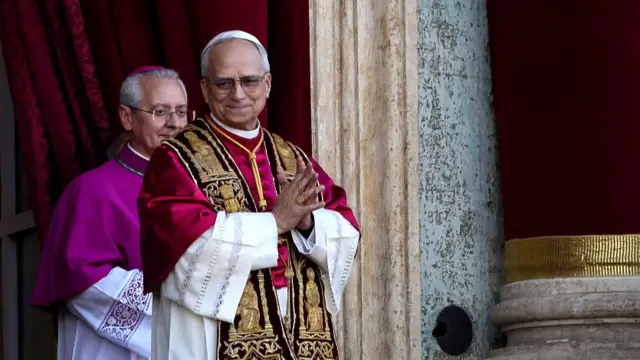 Pope Leo stands at the balcony in St Peter's Square. He is wearing red and gold robes. a priest in purple robes stands just behind him. Leo's hands are held in prayer at his chest.