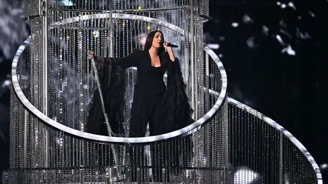 Yuval Raphael in black suit with very large long sleeves performs inside a light right