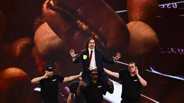 Tommy Cash performs on stage, a screen showing an image of a pile of coffee beans behind him