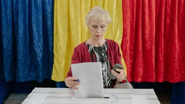 A woman (light blonde with thin frames resting on top of her head) in a red cardigan casts her vote into the ballot box. Behind her three voting booths closed by a blue (L), yellow (C) and red (R) curtain
