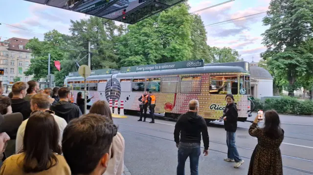 Eurovision fans watch the Eurovision karaoke tram go past as they queue