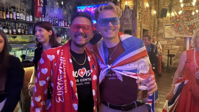 Two men adorned in Eurovision gear pose