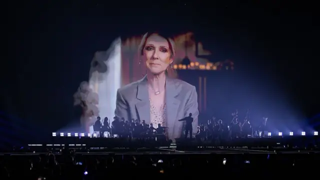 Video clip of Celine Dion in a grey blazer and cream blouse being played on screen as an orchestra plays at the bottom of it on the stage of the Eurovision semi-final