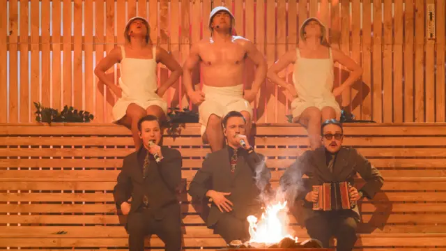 KAJ performing in a fake sauna with three dancers in towels behind them