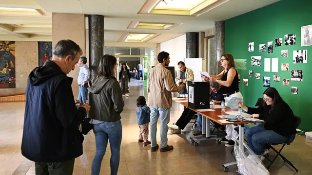 Voters queue to collect ballot papers at a public school in Lisbon, Portugal. At the front of the queue is a man holding hands with a toddler as he is handed a ballot paper