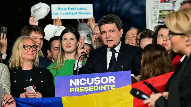 Nicusor Dan in a black suit addresses supporters and the press and he stands behind a podium bearing a sign reading Nicusor President, a Romanian flag in the bottom of the frame