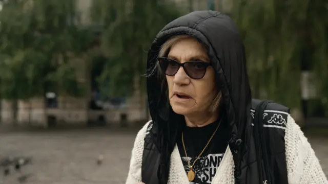 A woman with her mouth partially open, wearing a cardigan and hoody and dark glasses