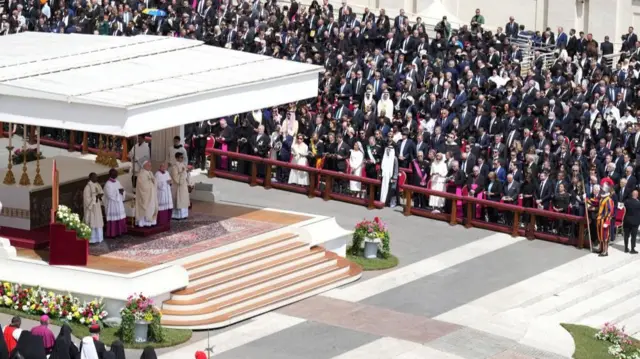 A landscape shot of the Pope with others dressed in religious clothing standing in the shade of an overhead shelter. Those attending are seated to the left and right of the platform where he stands.