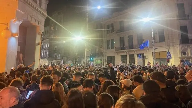 Large crowd of people in a dark street, a couple of EU flags being flown on high poles as people hold up their phones