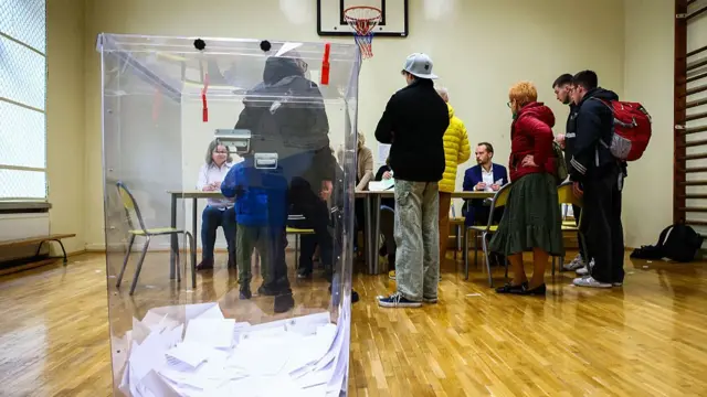 A group of people wait in front of a table to collect polling cards in a school gym turned into a polling station in Poland