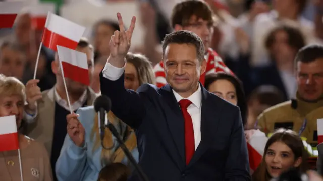 Rafal Trzaskowski hold his right hand up in a victory sign as supporters applaud him at the back waving Polish flags