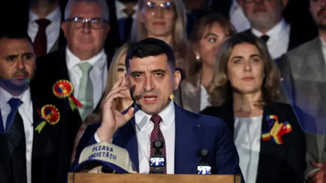 George Simion in dark blue suit addresses supporters outside of Romanian parliament after exit polls. Behind him are supporters wearing rosettes in the colours of the Romanian flag (Blue, yellow and red)