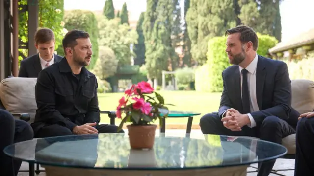 Zelensky and JD vance sat on table with garden in background.