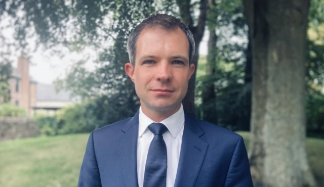 Andrew Bowie wearing a blue suit looks directly at the camera against the backdrop of some trees and greenary