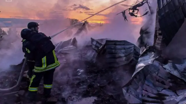 Two firefighters in black and yellow outfits, pointing hose with water at building with burnt metal nearby on the ground.