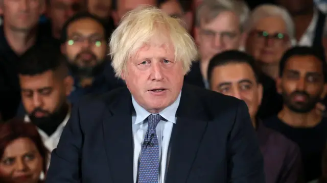 Boris Johnson wears a suit. he is speaking at an event and people can be seen sitting in the audience behind him. It is a close cropped image of his head and shoulders.
