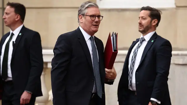 Starmer in a suit holding a red folder, outdoors, stone wall in background