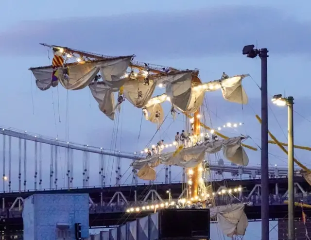 Sailors in harnesses hanging off the masts of the ship as it sits in the river