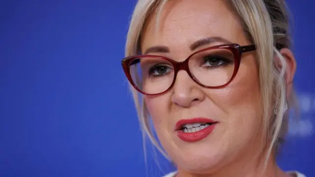 Michelle O'Niell, close up of face against a blue background. She is mid-speech, indoors, wearing glasses.