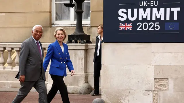 Antonio Costa is in a grey suit, and von der Leyen in a bright blue blazer. Both are outside, mid-stride against a stone wall backdrop