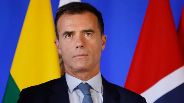 Photograph of Sandro Gozi, chair of the EU-UK Parliamentary Partnership Assembly, wearing a blue tie, suit and blue shirt with a backdrop of some flags behind him
