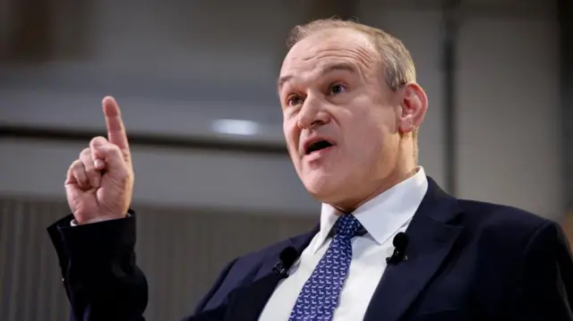 Ed Davey speaking indoors, holding a finger in the air.