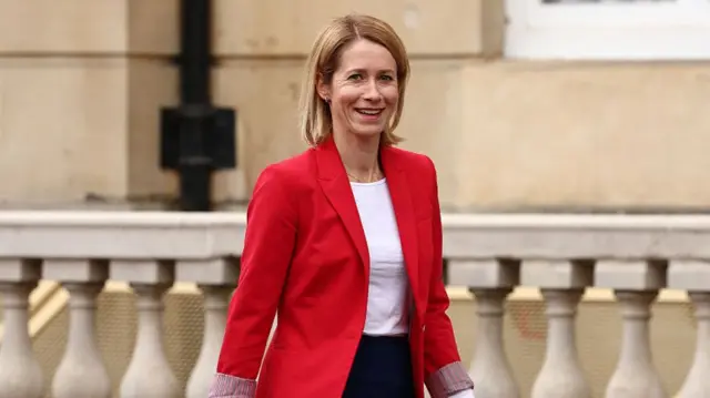 Kallas is wearing a bright red blazer and a white top. She is outside, in front of a stone building