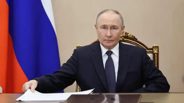Putin in a chair with Russian flag behind him.
