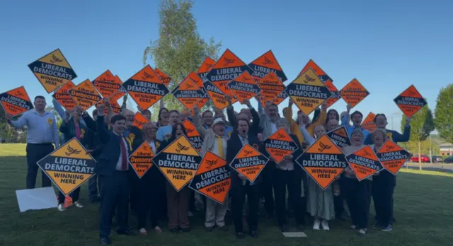A group of lib dems on a green lawn with a blue sky. they are cheering and holding up orange diamond shaped lib dem signs.